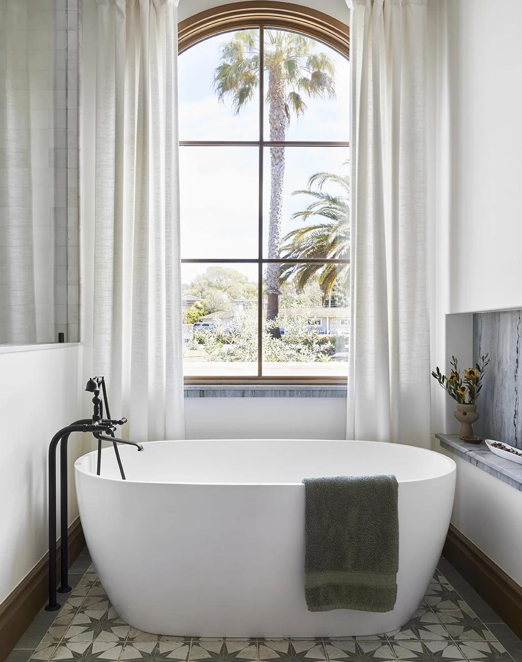 Bright bathroom with a freestanding tub and an arches window behind. Green and Cream mosaic on the floor and a blue marble slab nook on the right of the tub.